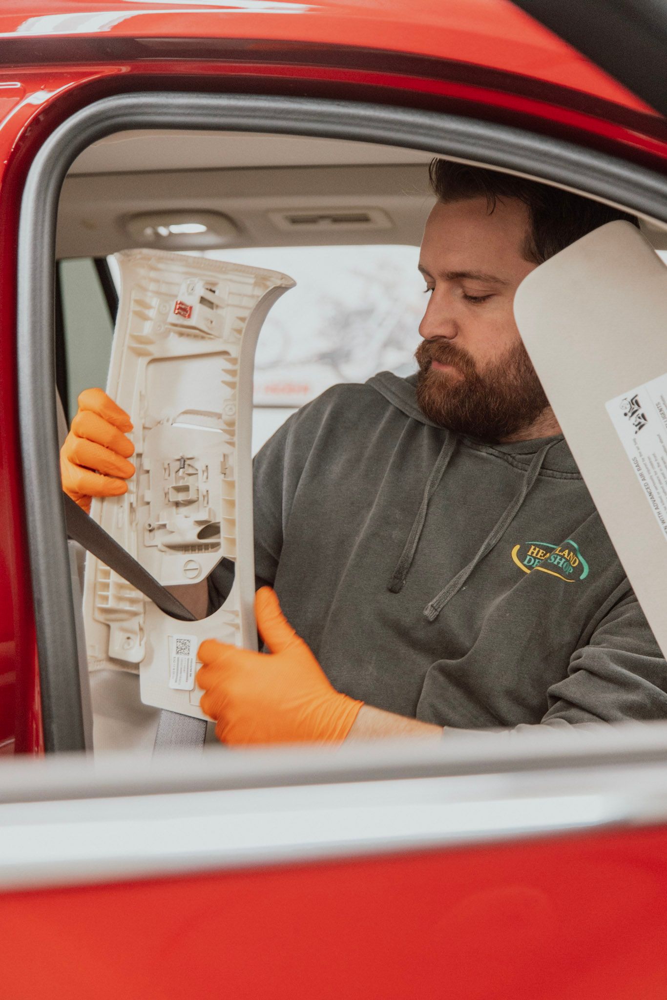 Man in orange gloves works on a car, holding a white part, looking inside. Red car door.