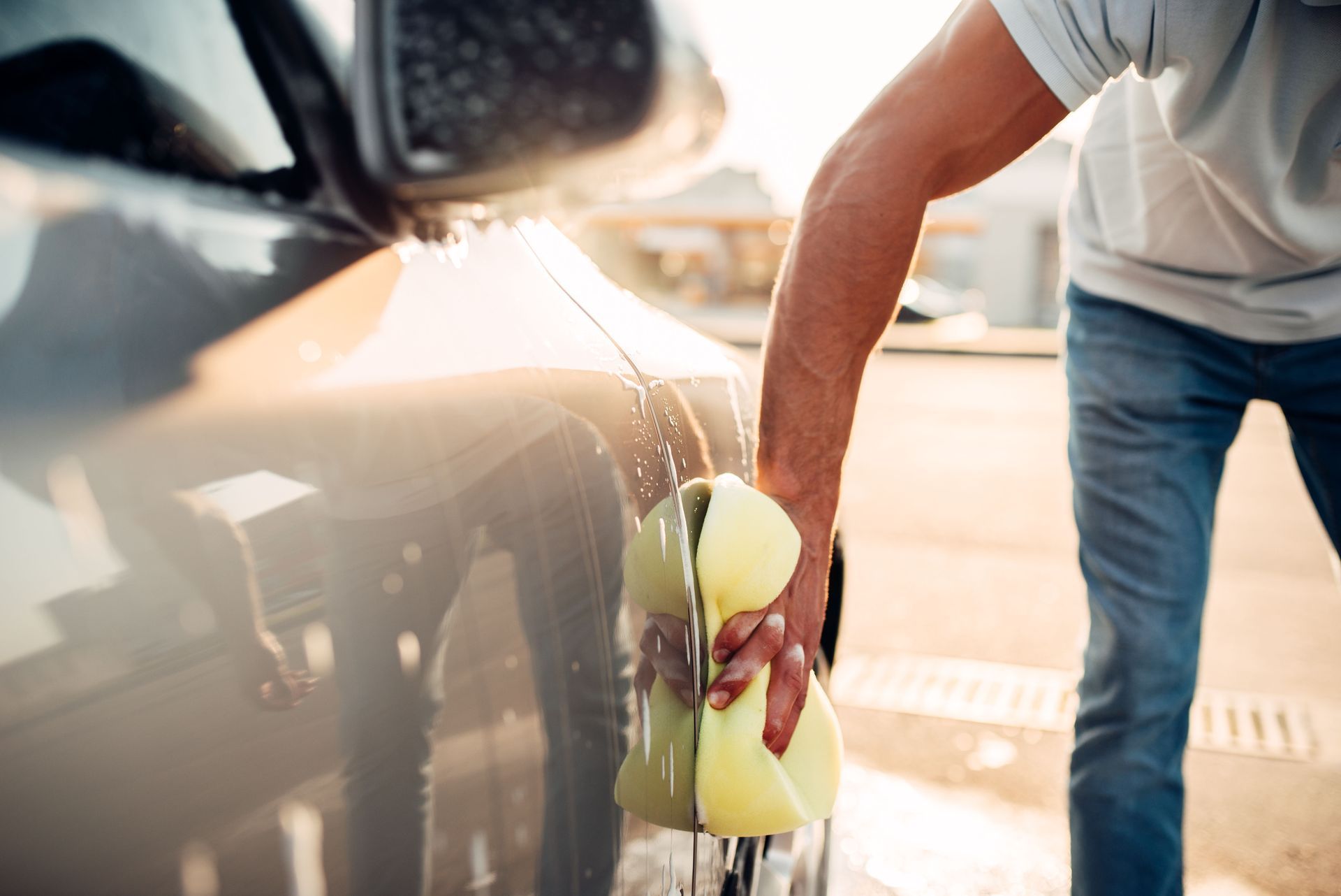 Person washing a car with a sponge. Water and suds visible. Sunny outdoor setting.