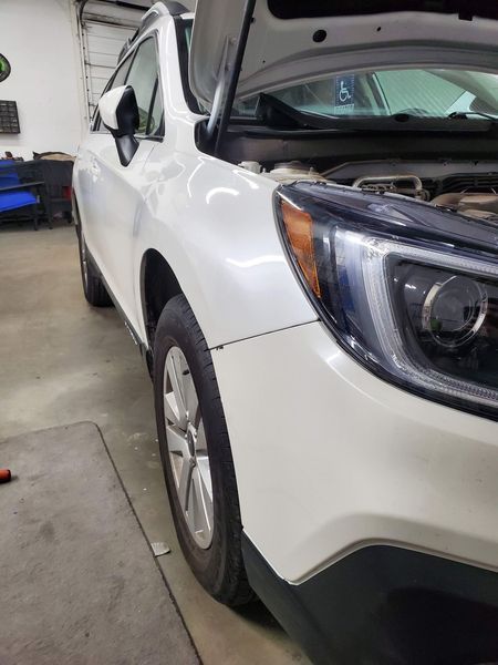 White Subaru Outback car with hood open in a garage, showcasing engine components.