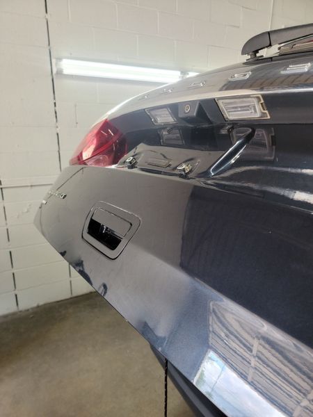 Rear view of a dark gray car's trunk, showing a taillight, license plate area, and handle, likely in a garage.