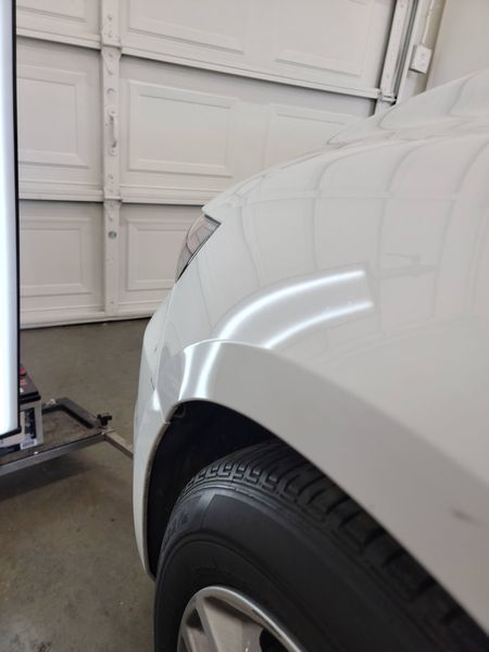 White car front fender with a dent, tire, and garage door in background.