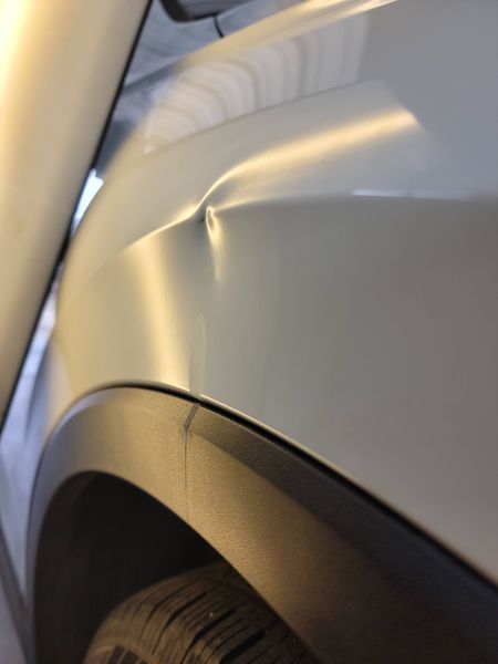 Silver car fender with a large dent above the wheel well, near the door.