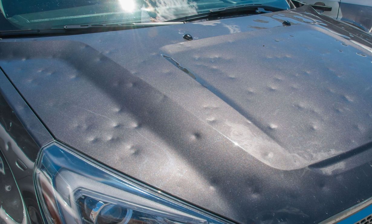 Gray car hood heavily dented, likely from hail damage. Sun glares off the surface.