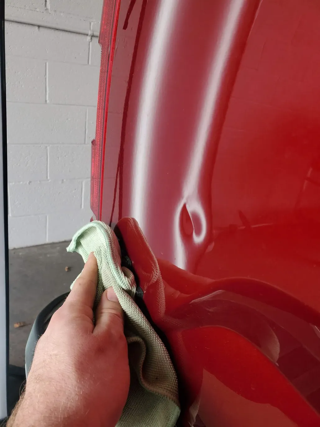 Person's hand wiping a red car's body with a green cloth, focusing on a dent. Garage setting.