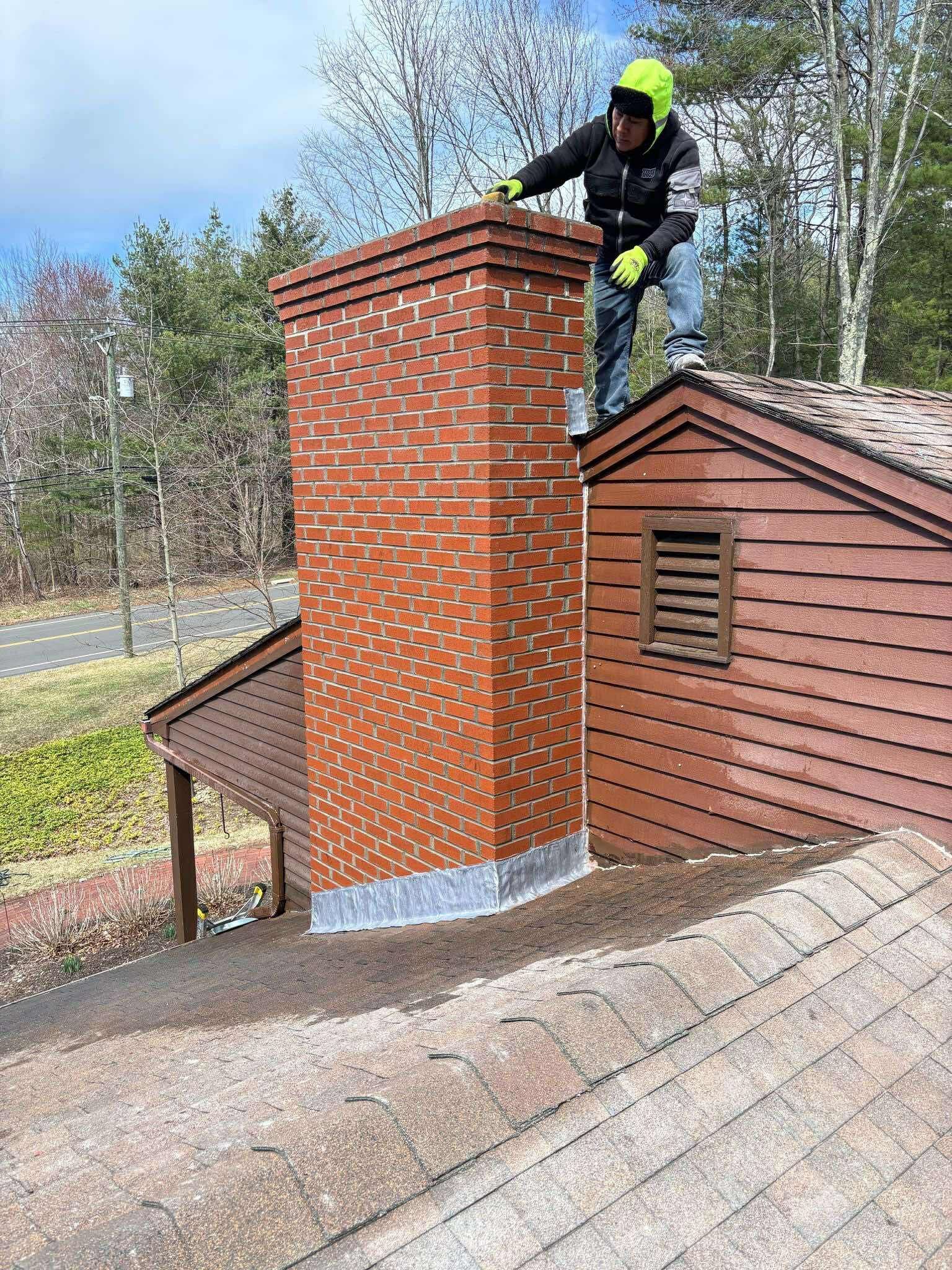 A person on a roof repairs a tall brick chimney; brown shingles, green trees and blue sky.