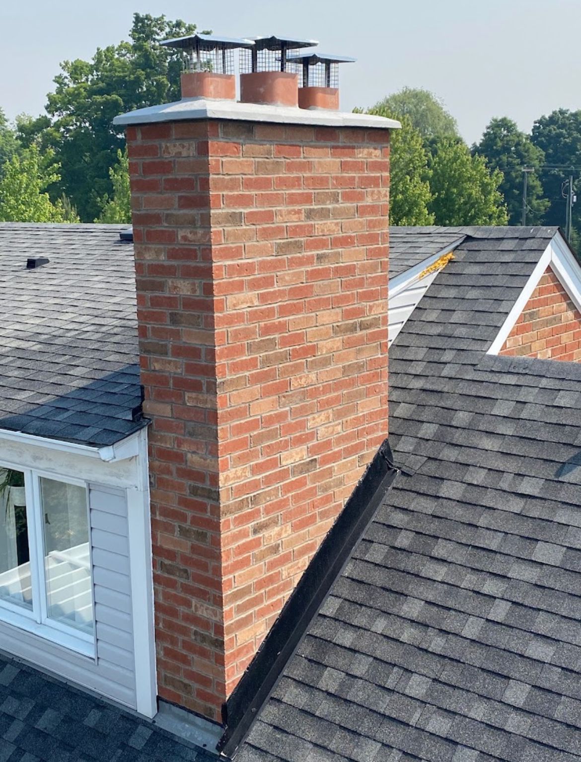 Brick chimney on a roof, with three chimney caps, set against a backdrop of trees and sky.