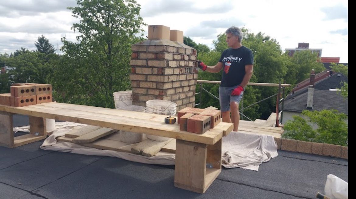 Man building a brick chimney on a rooftop with a wooden work platform, trees in the background.