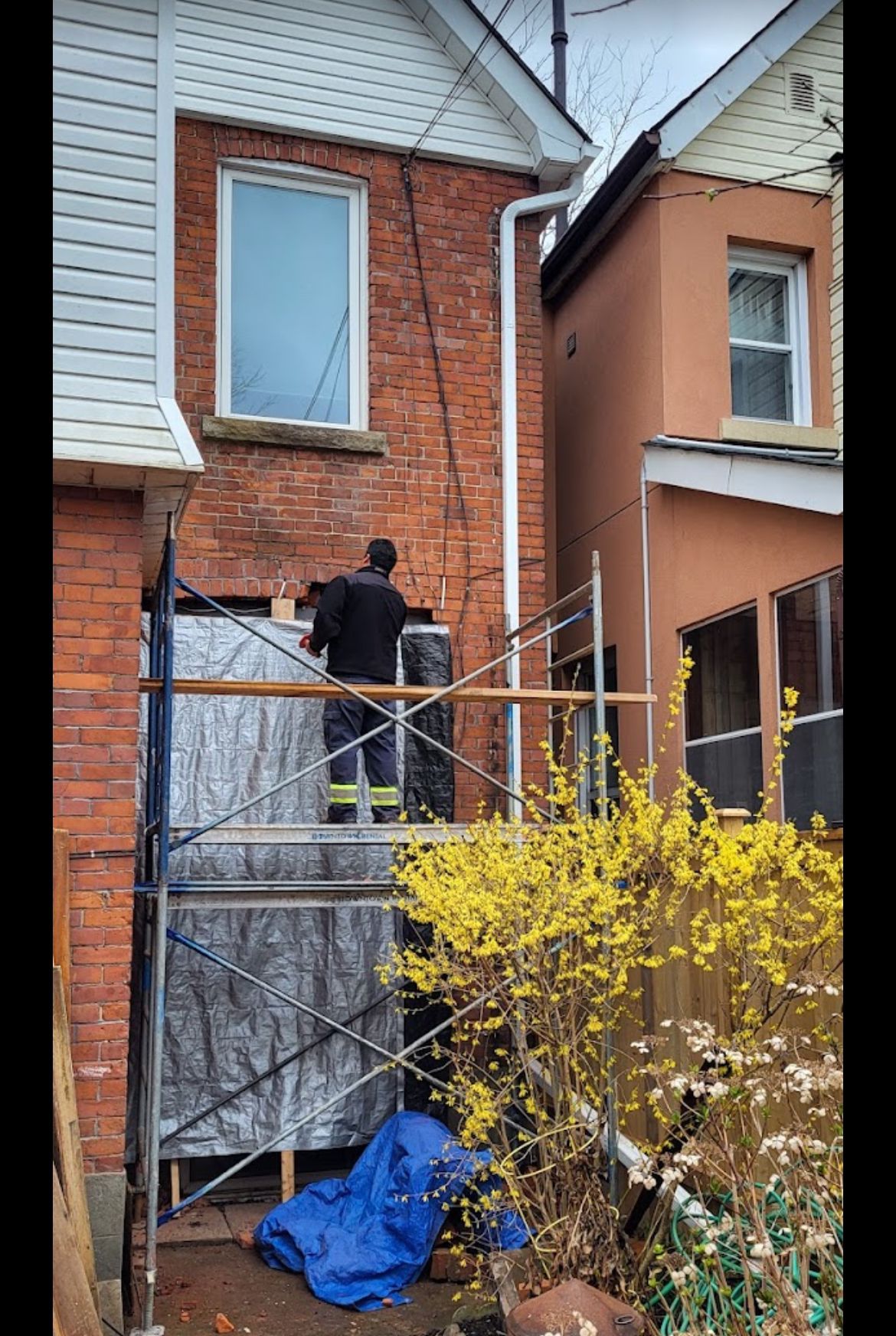 A person on scaffolding repairs a brick house exterior. Yellow flowers bloom below.