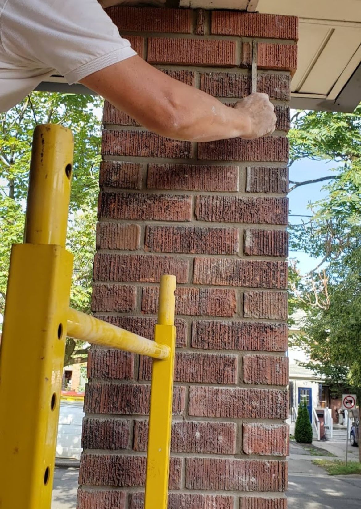 Person using a tool on a brick pillar, supported by a yellow scaffold; outdoors.