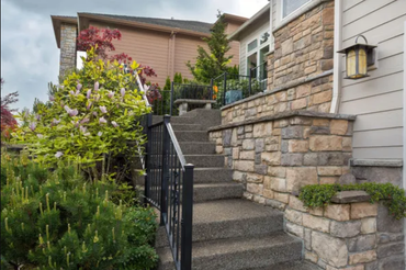 Stone steps lead to a house entrance, flanked by black railing and lush green and red foliage under a cloudy sky.