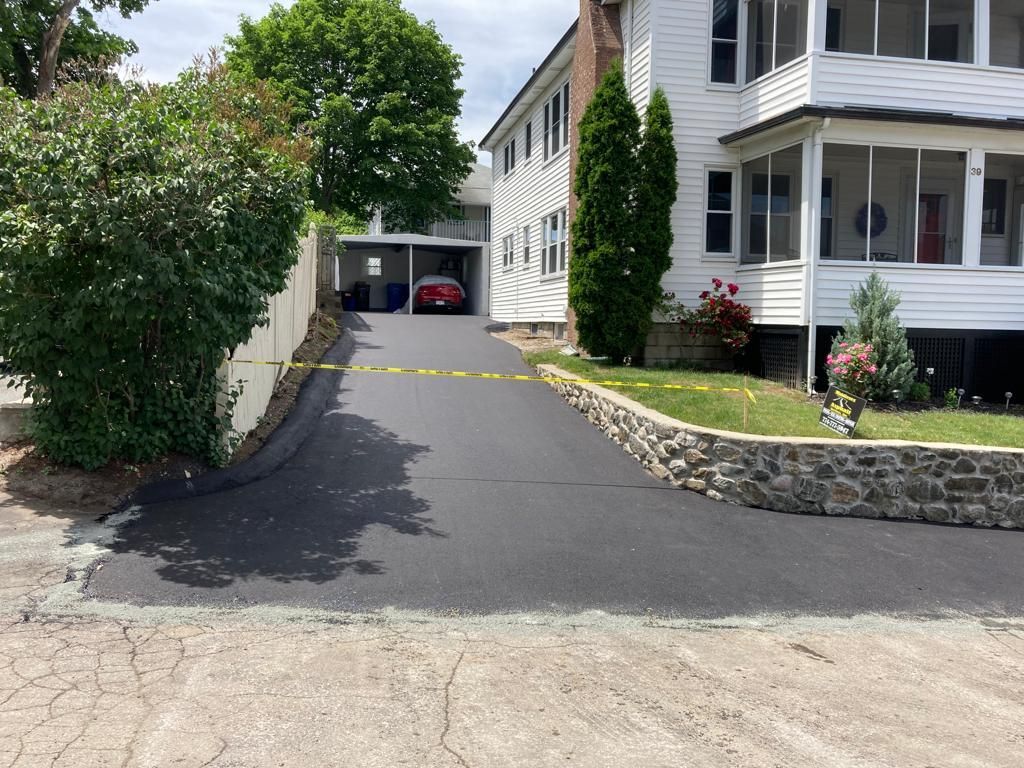 Newly paved driveway leading to a white house with a red car in a carport, surrounded by greenery.
