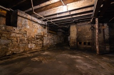 Dark, aged basement with exposed stone walls, wooden beams, and a concrete floor.