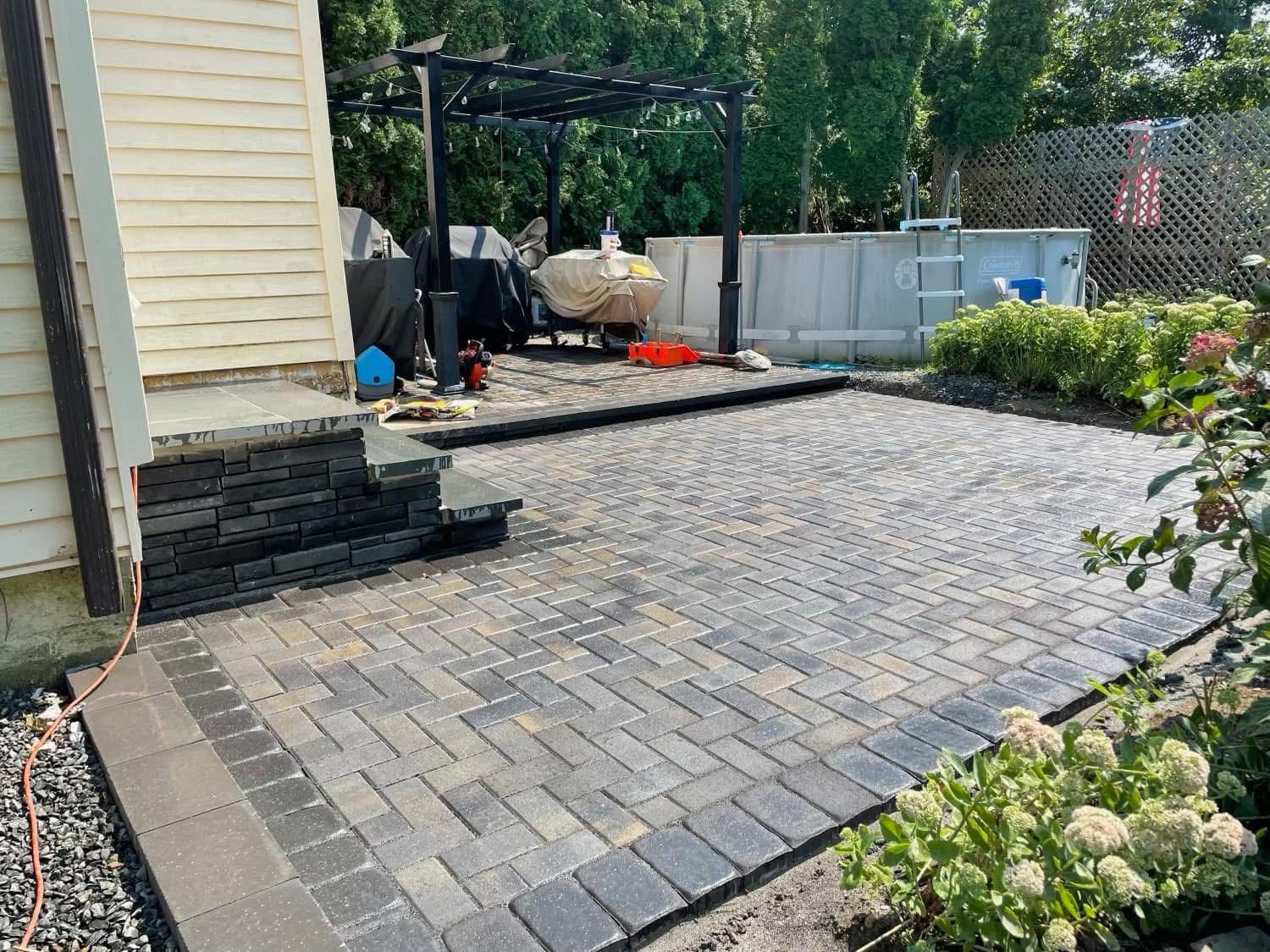 Stone patio with steps next to a house. A pergola, pool, and plants are in the background.