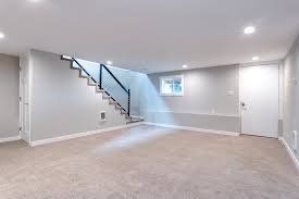 Empty, light gray basement room with stairs, carpeted floor, and a white door.