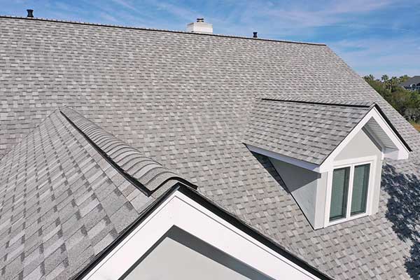 Gray shingled roof of a house with a dormer window against a blue sky.