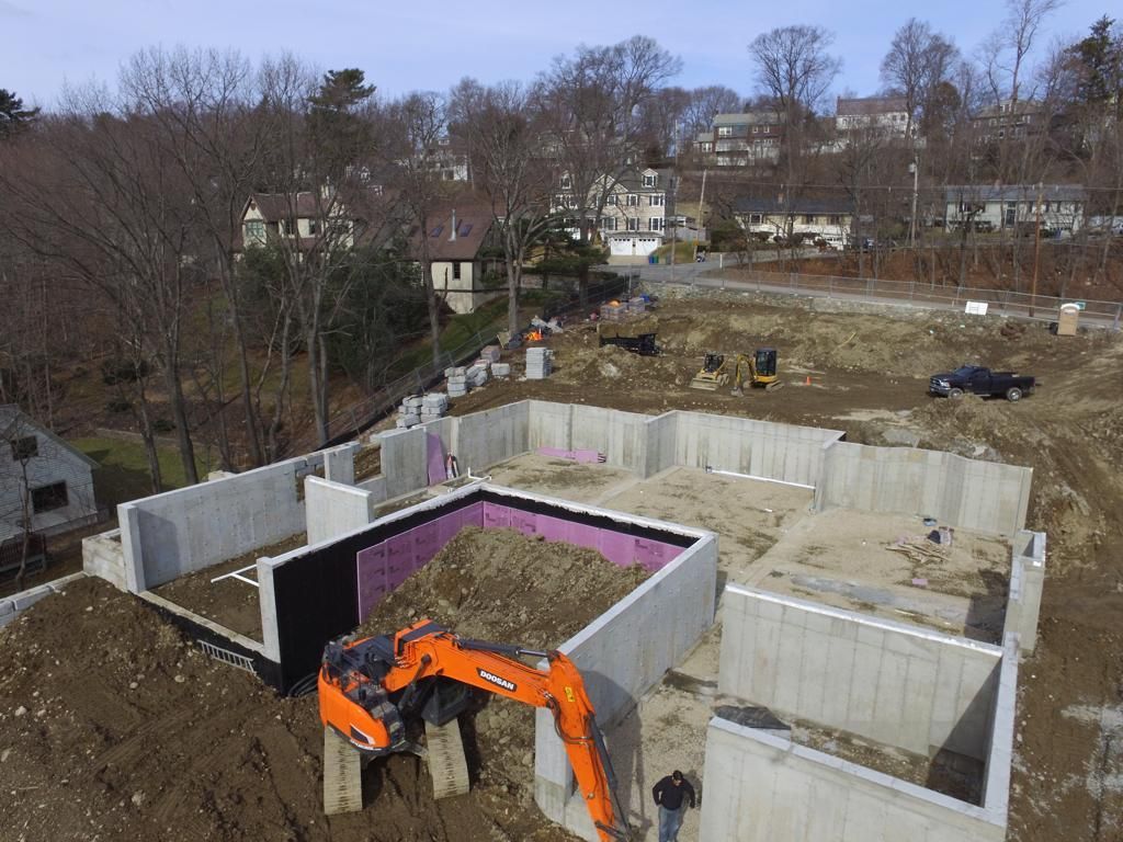 Excavator digging at a building's foundation site, with a house under construction and other houses in the background.