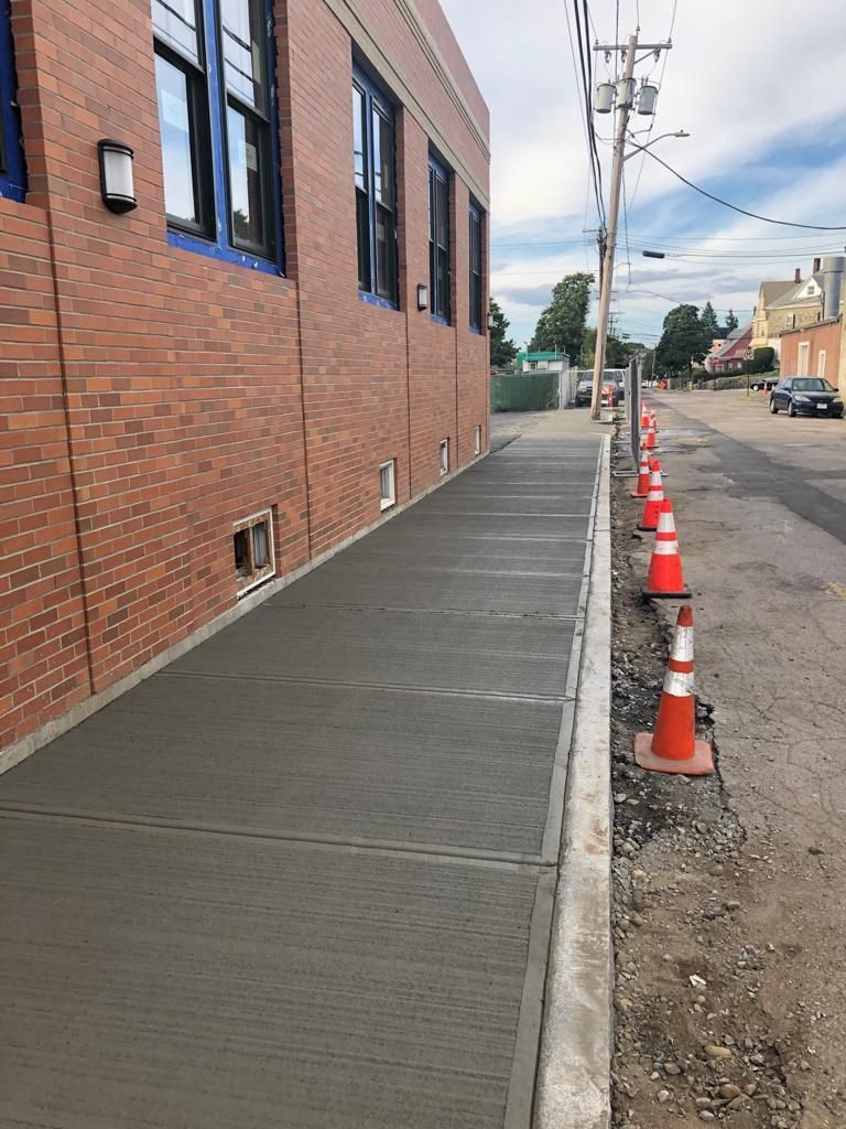 Freshly poured concrete sidewalk alongside a brick building, with orange traffic cones lining the street.