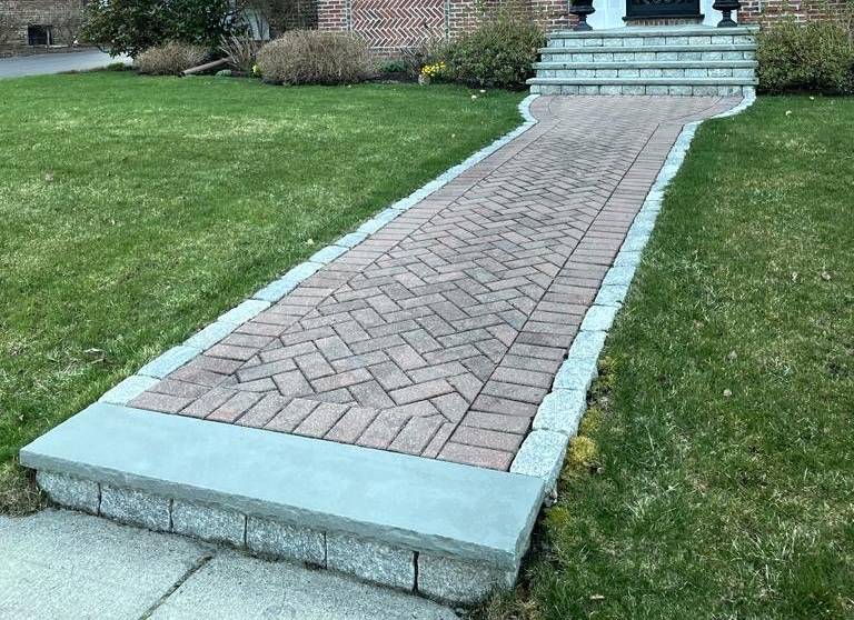 Brick walkway leading to a house with steps, bordered by grass and stone edging.