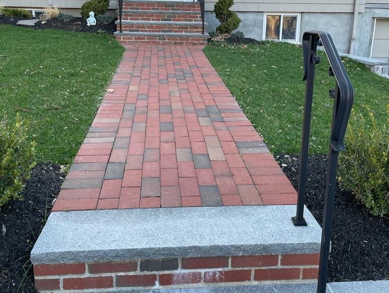 Brick walkway leading to steps, flanked by green grass and landscaping. Railing on the right.