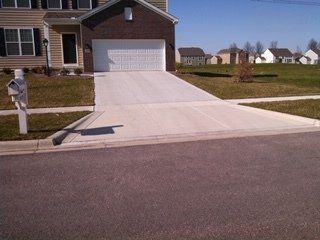 Concrete driveway leading to a house with a two-car garage. Curb and road in foreground, grass and other houses in background.