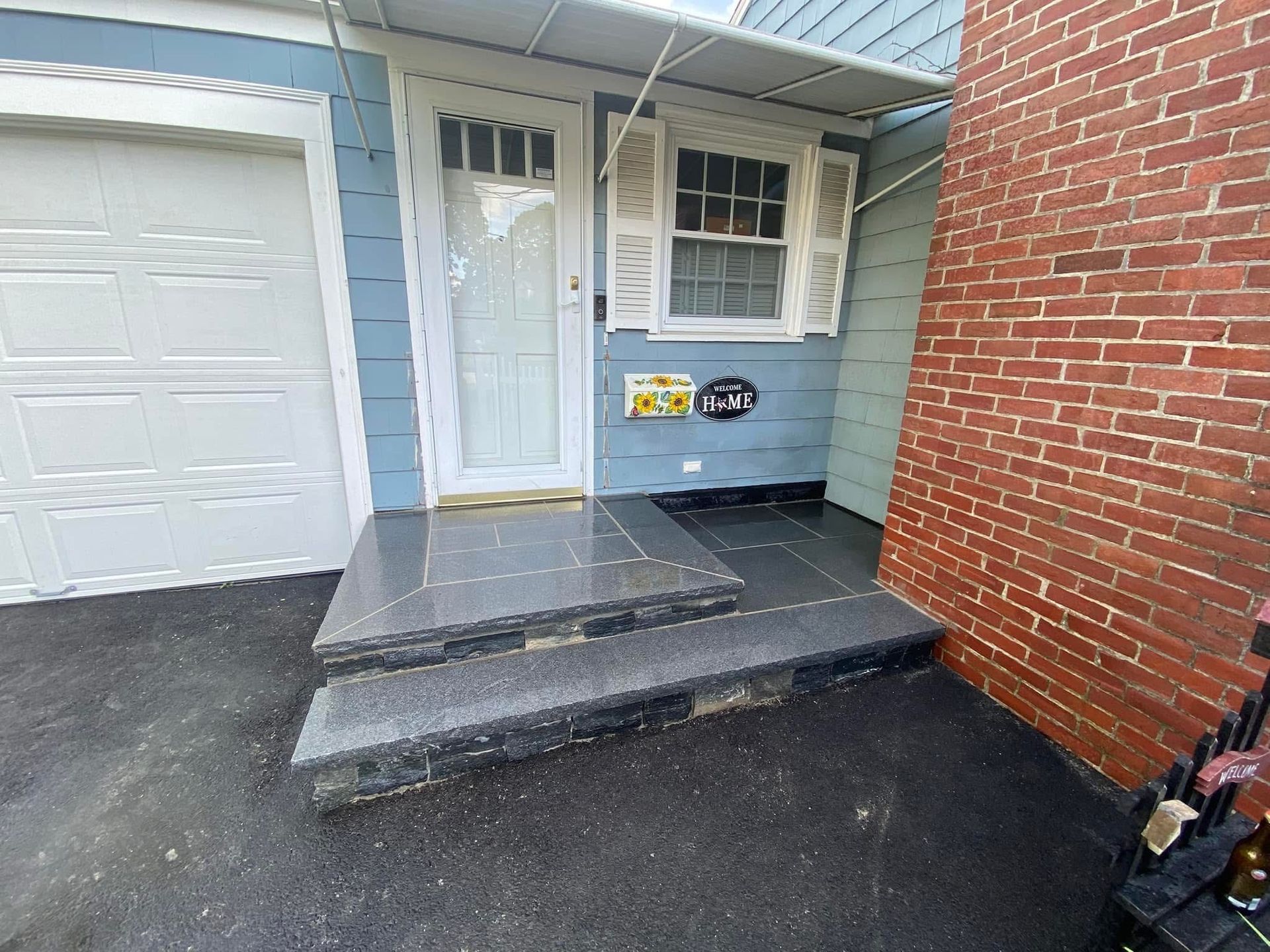 Blue house entrance with gray steps and a white garage door, next to a red brick wall.