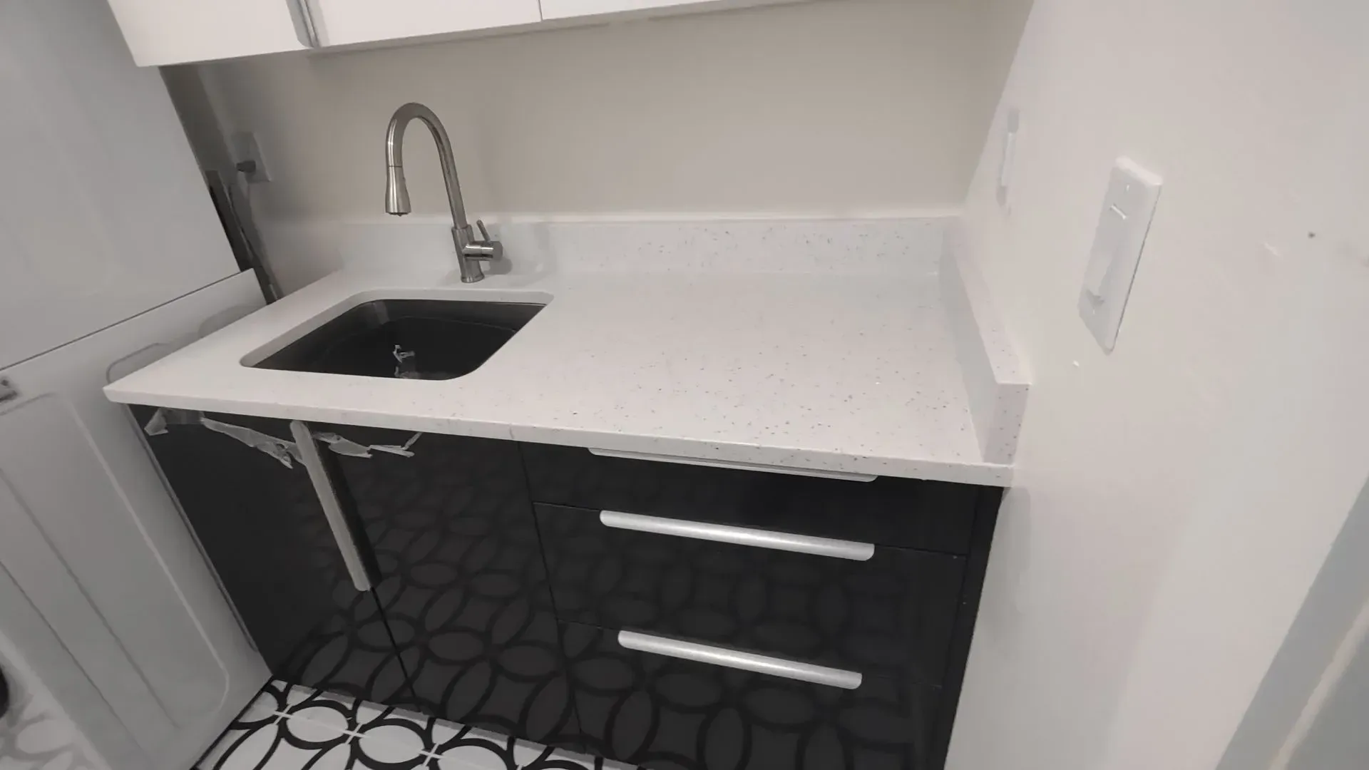 Small kitchen counter with black sink, chrome faucet, white countertop, and dark drawers on patterned floor.