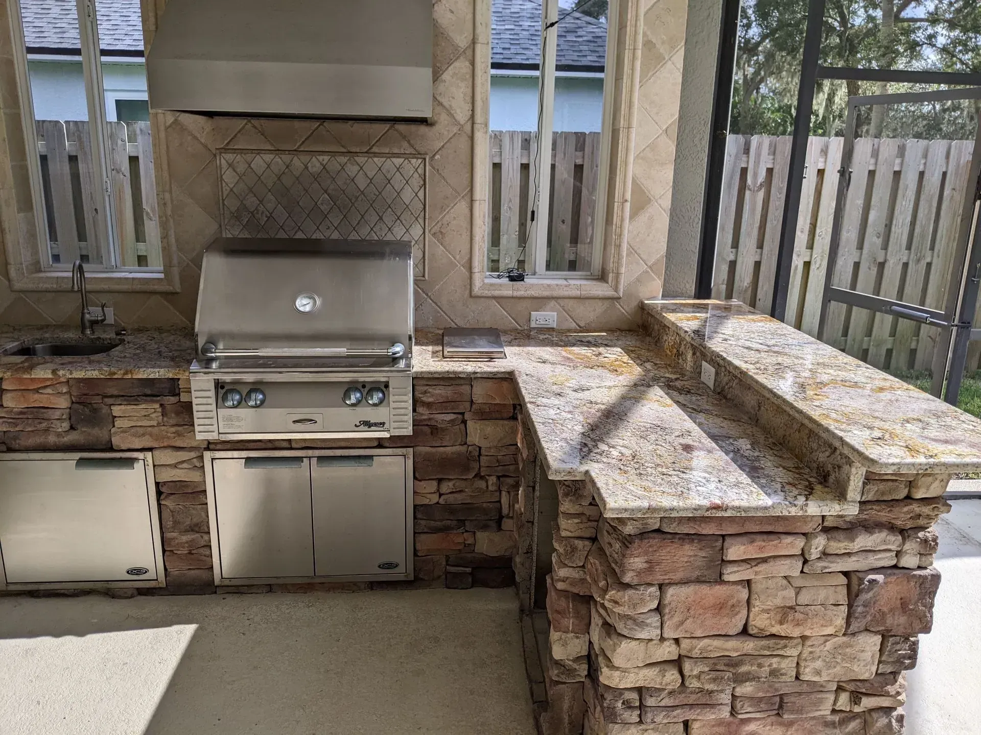 Outdoor stone patio kitchen with stainless grill, sink, and beige countertops by a fence
