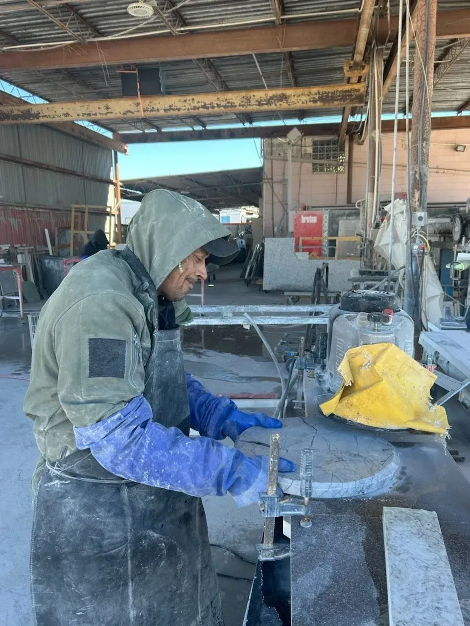 Worker in hooded jacket using a power tool in a dusty workshop, with a yellow bag nearby.