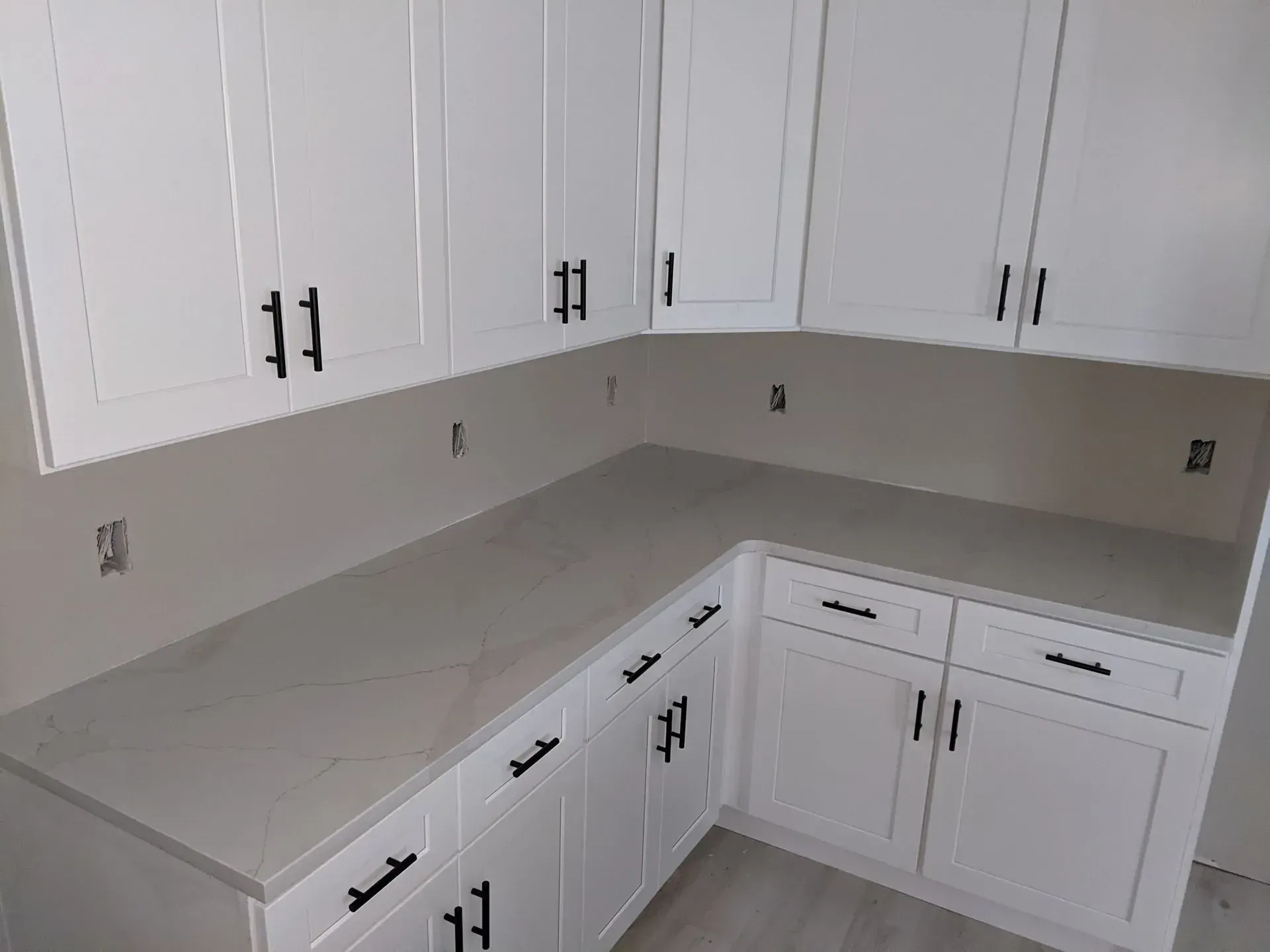 White L-shaped kitchen with upper and lower cabinets, gray countertop, and beige backsplash.