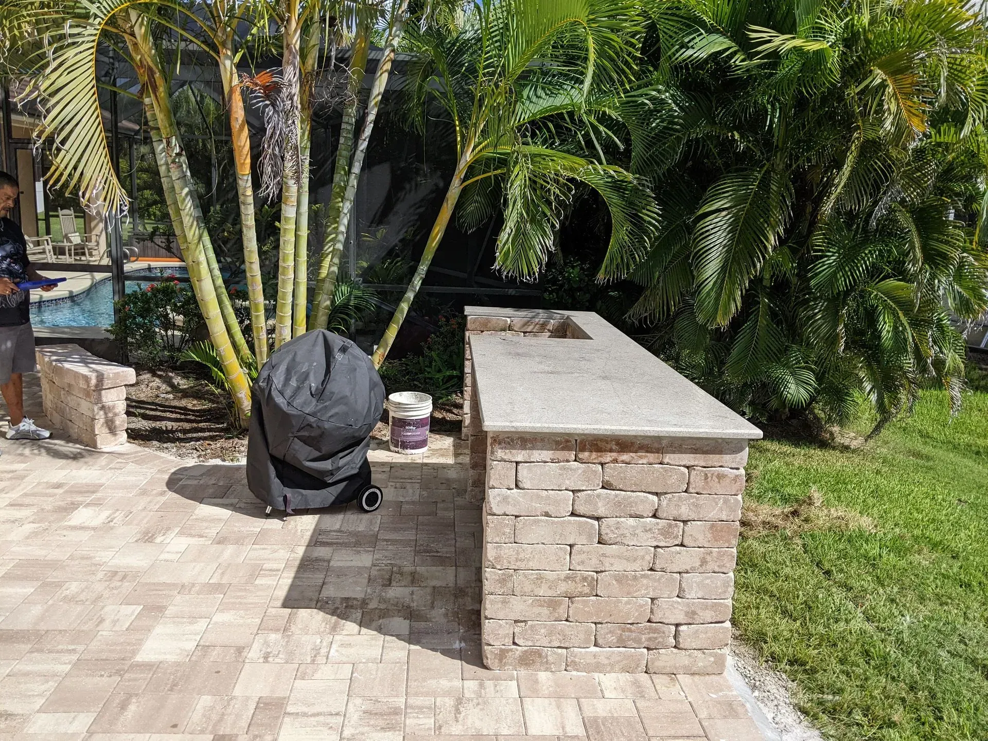 Backyard patio with a brick bench, potted plants, and a black-covered object beside a grassy lawn.