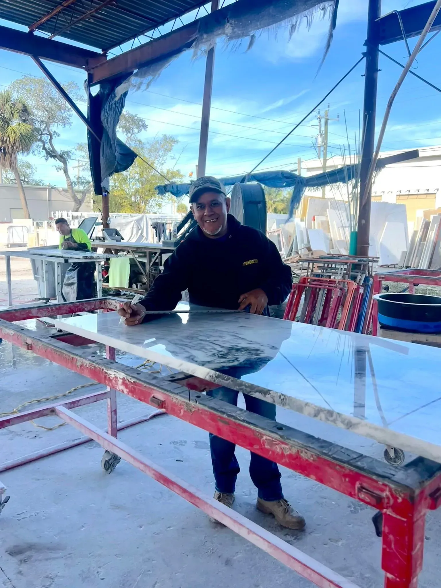 Person in a black coat standing beside a large table under a covered outdoor workspace.