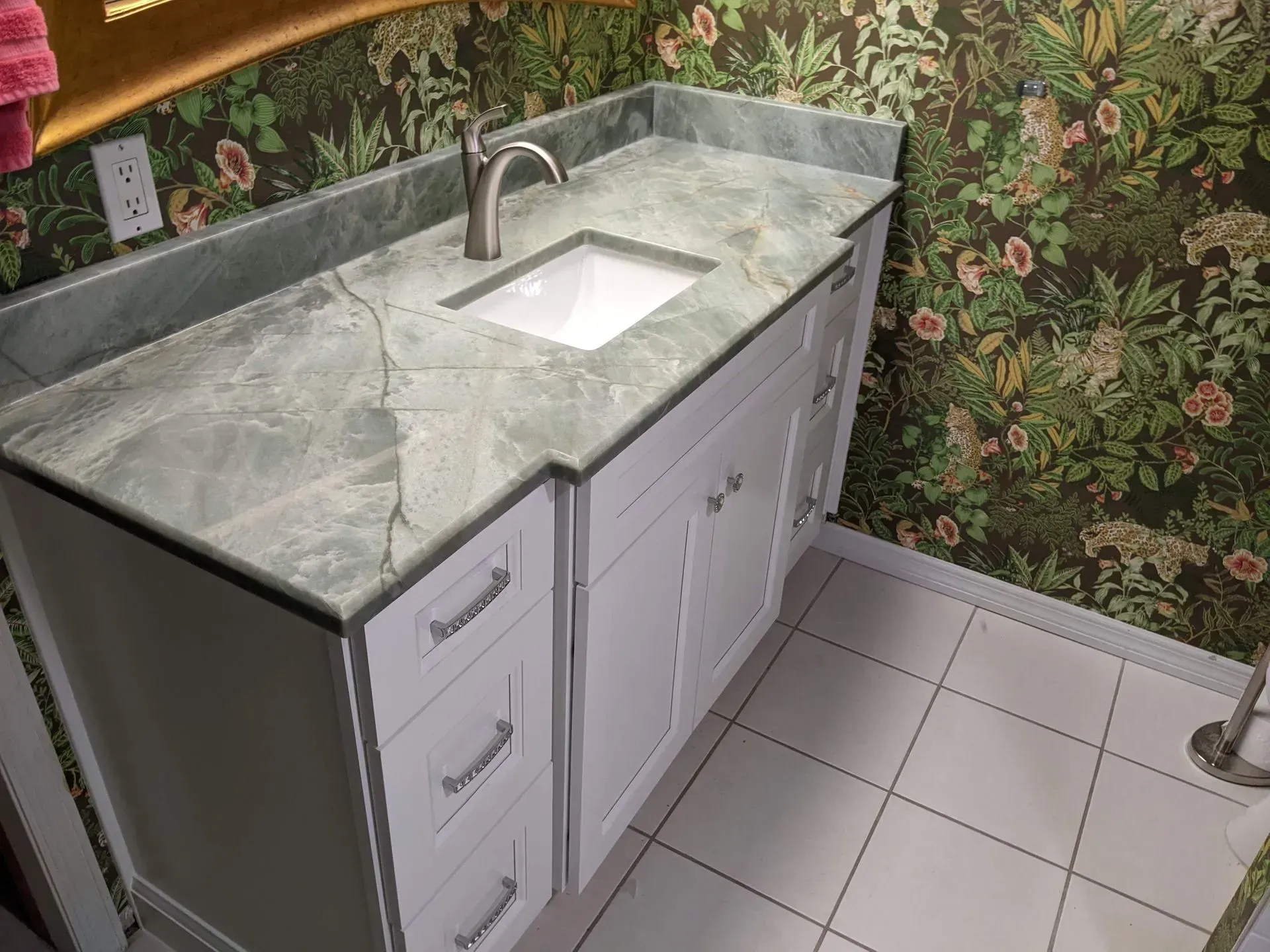 White bathroom vanity with marble countertop, sink, and floral wallpaper in a tiled room