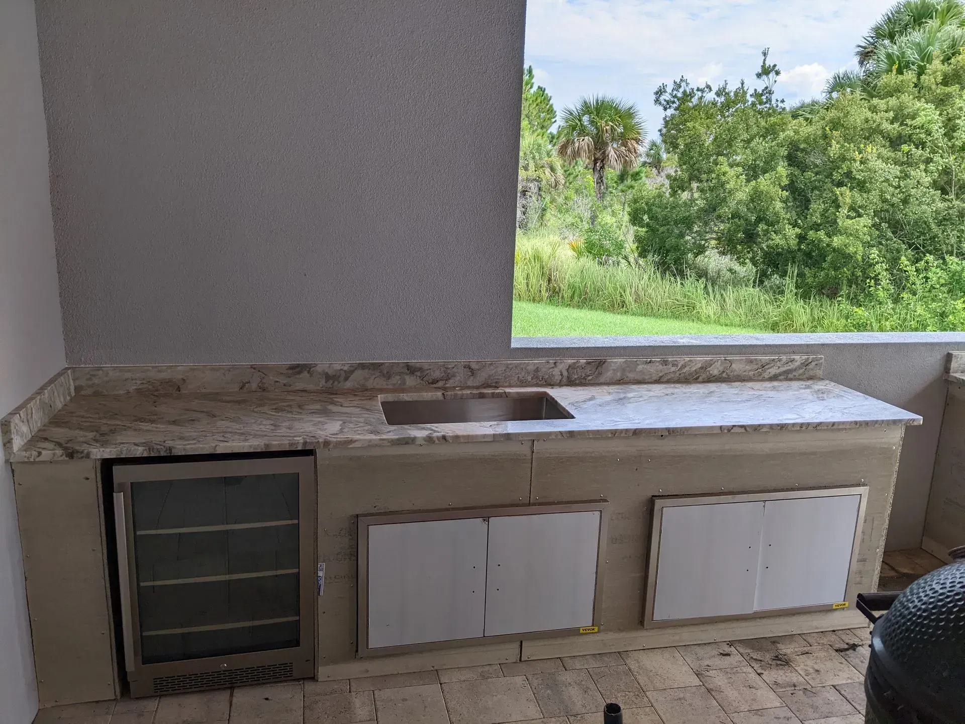 Outdoor stone countertop with sink, cabinets, and a grill under a covered patio, overlooking greenery