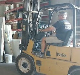 Worker driving a yellow Yale forklift in a warehouse aisle beside shelves and stacked goods