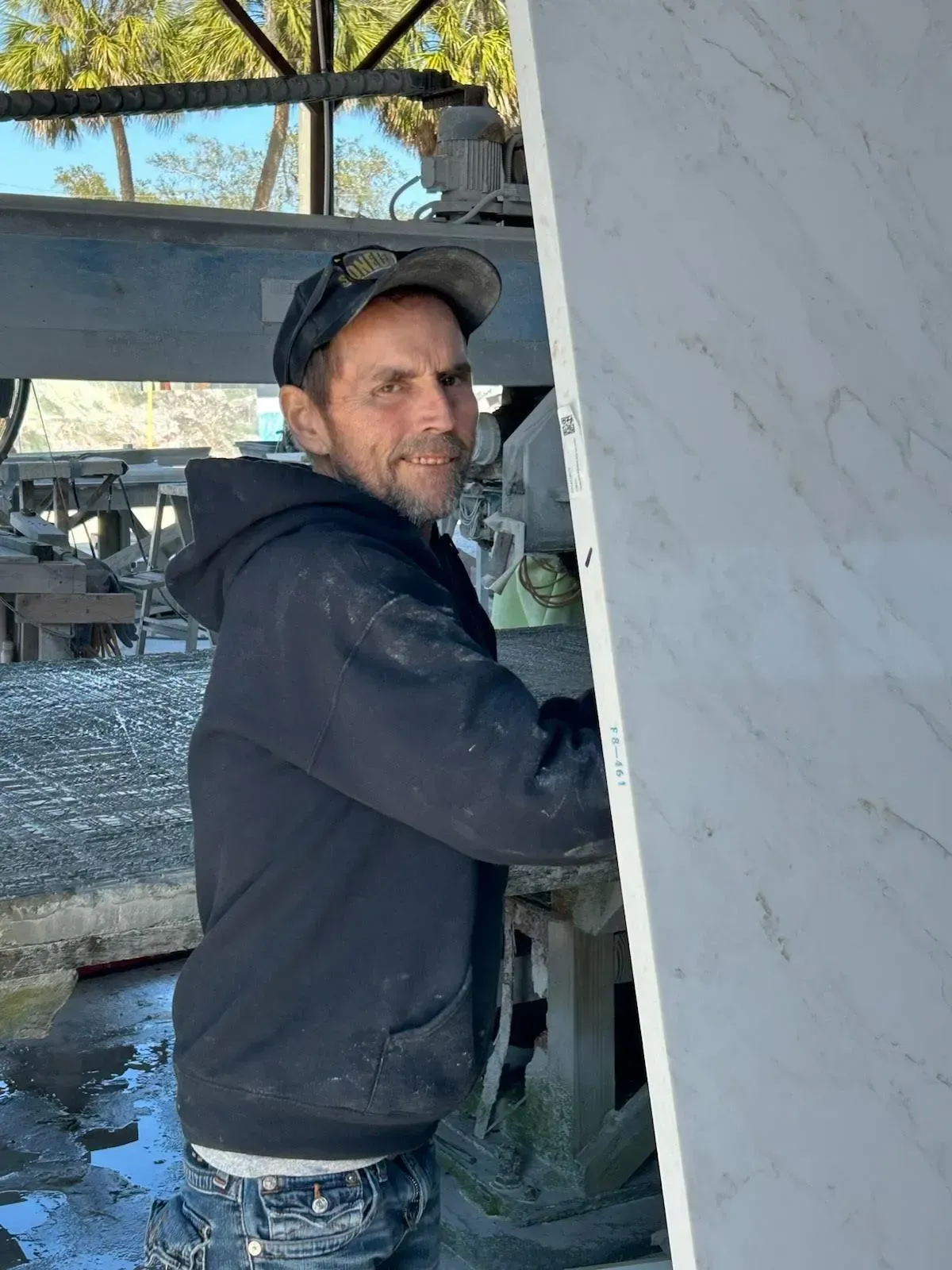 Worker in a dark hoodie and cap standing beside large marble slabs outdoors.