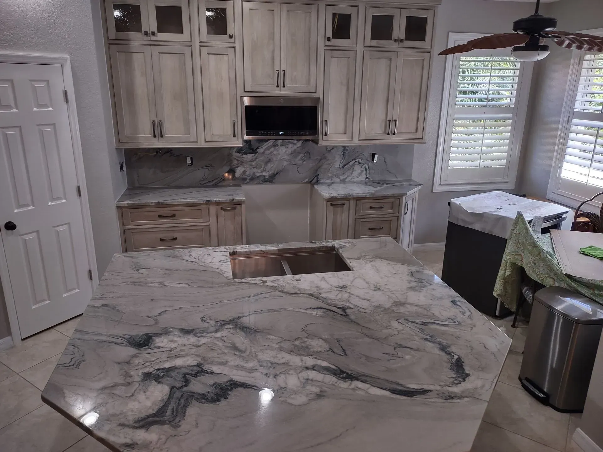 Bright kitchen with white cabinets, marble countertops, central island, and ceiling fan near shuttered window.