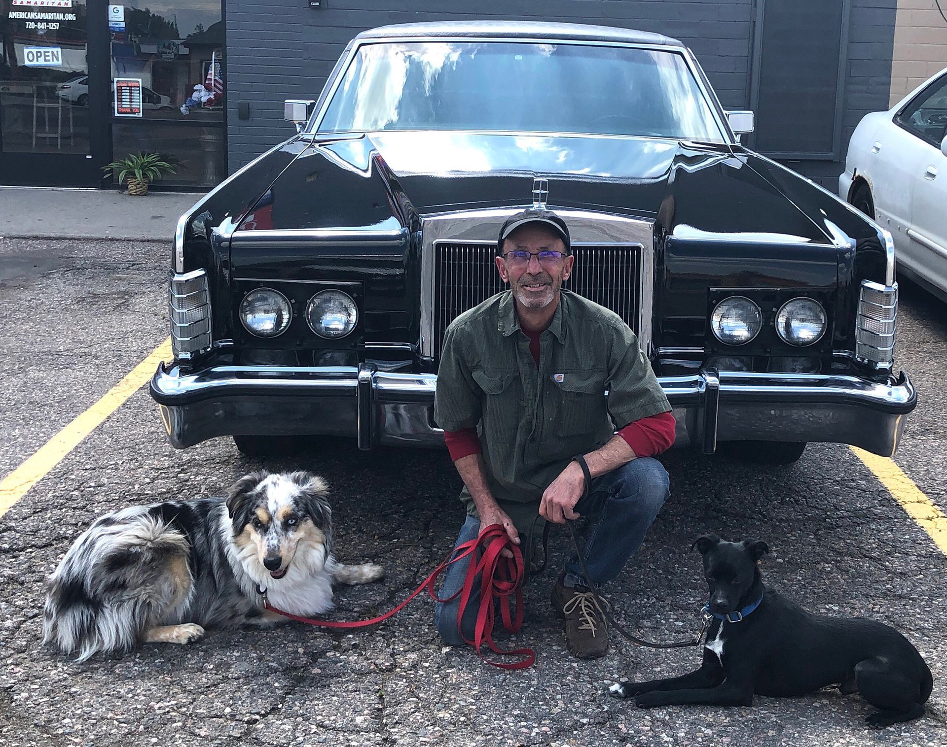 A man is kneeling down in front of a car with two dogs.