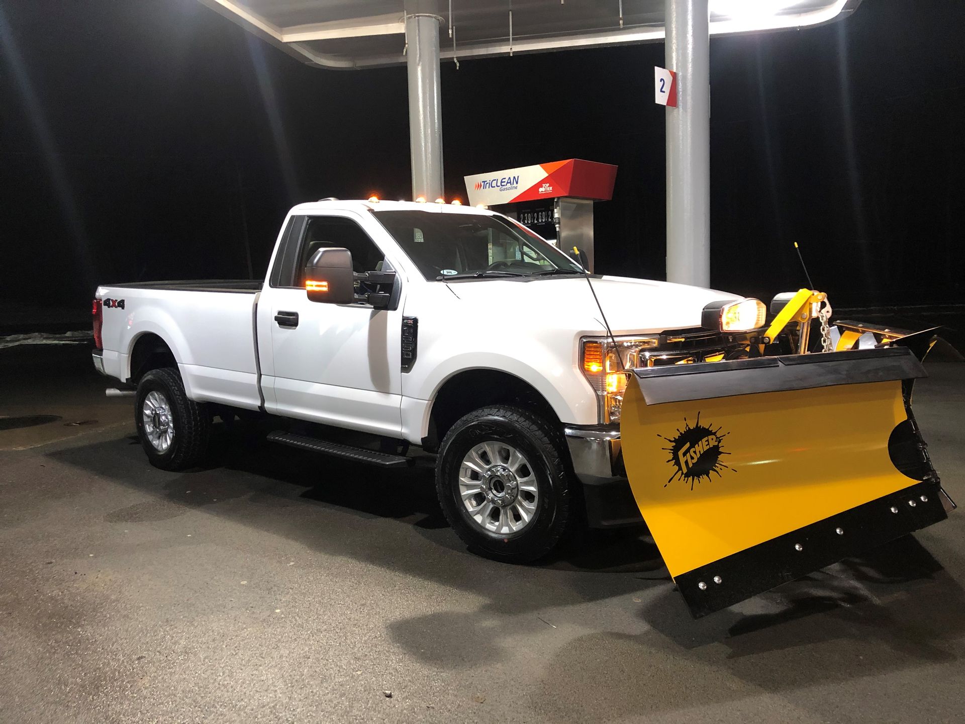A white truck with a yellow snow plow attached to it is parked at a gas station.