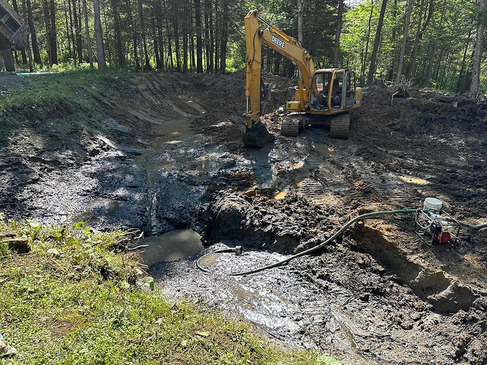 A yellow excavator is digging a hole in the mud in the middle of a forest.