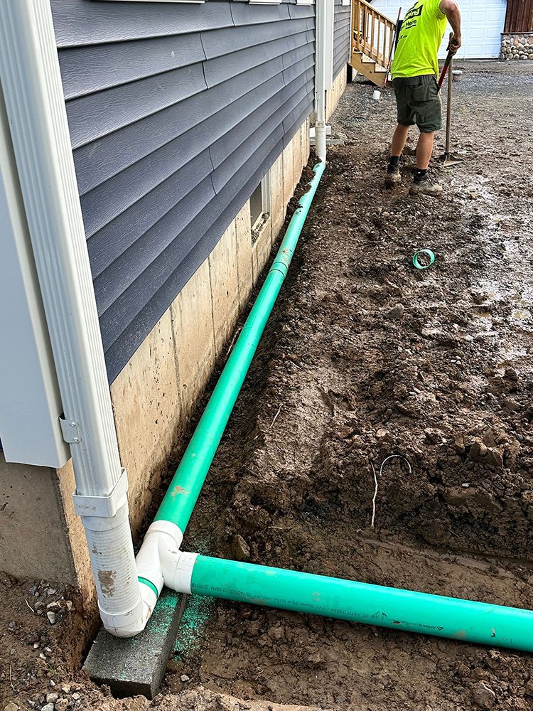A man is digging in the dirt next to a house with green pipes.