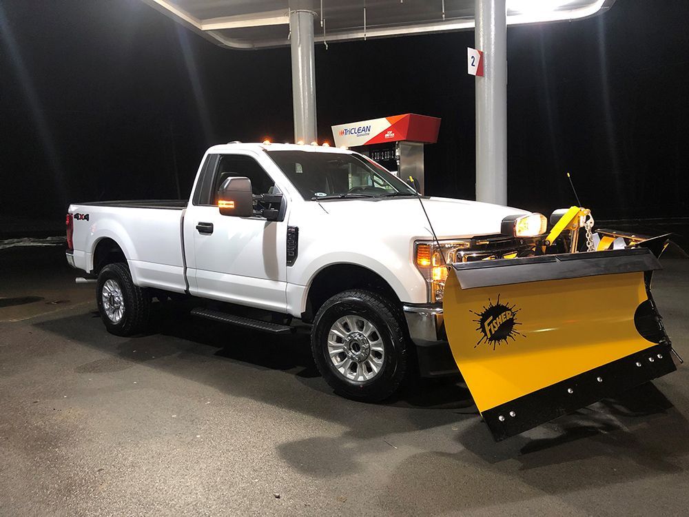 A white truck with a yellow snow plow is parked at a gas station.