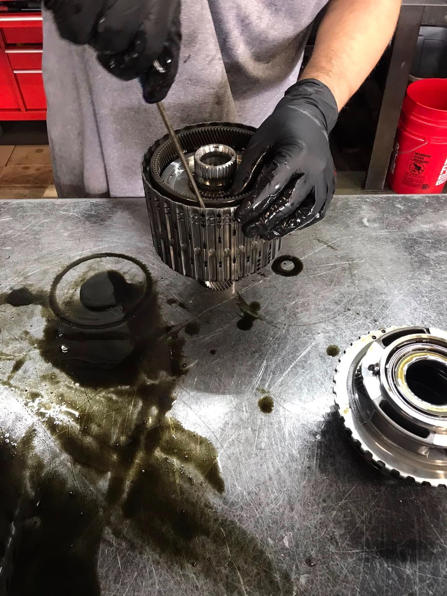 Person with black gloves working on a car part on a workbench, with spilled oil nearby.
