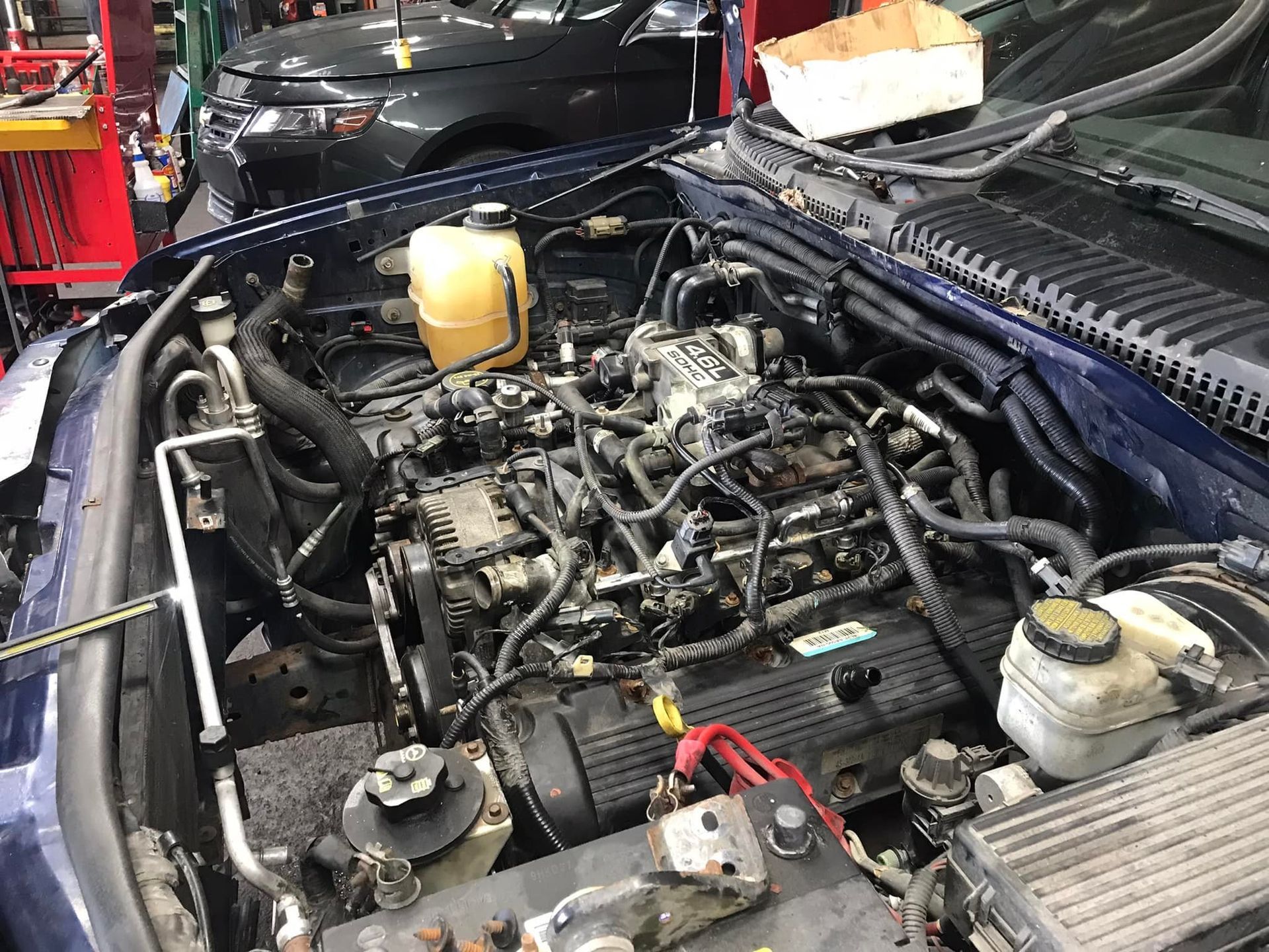 Blue Ford Explorer engine bay, being serviced in a garage. Dark engine with hoses and belts visible.