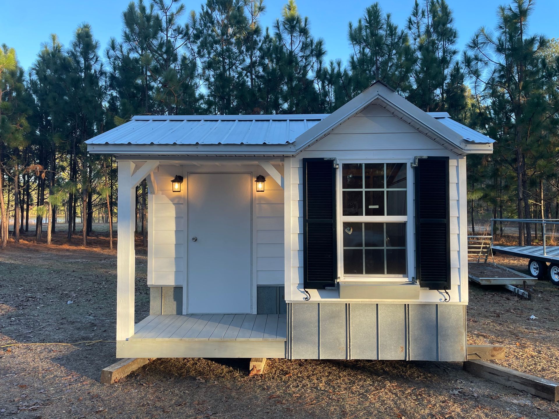 Small white shed with porch, door, and window in a wooded outdoor setting at dusk