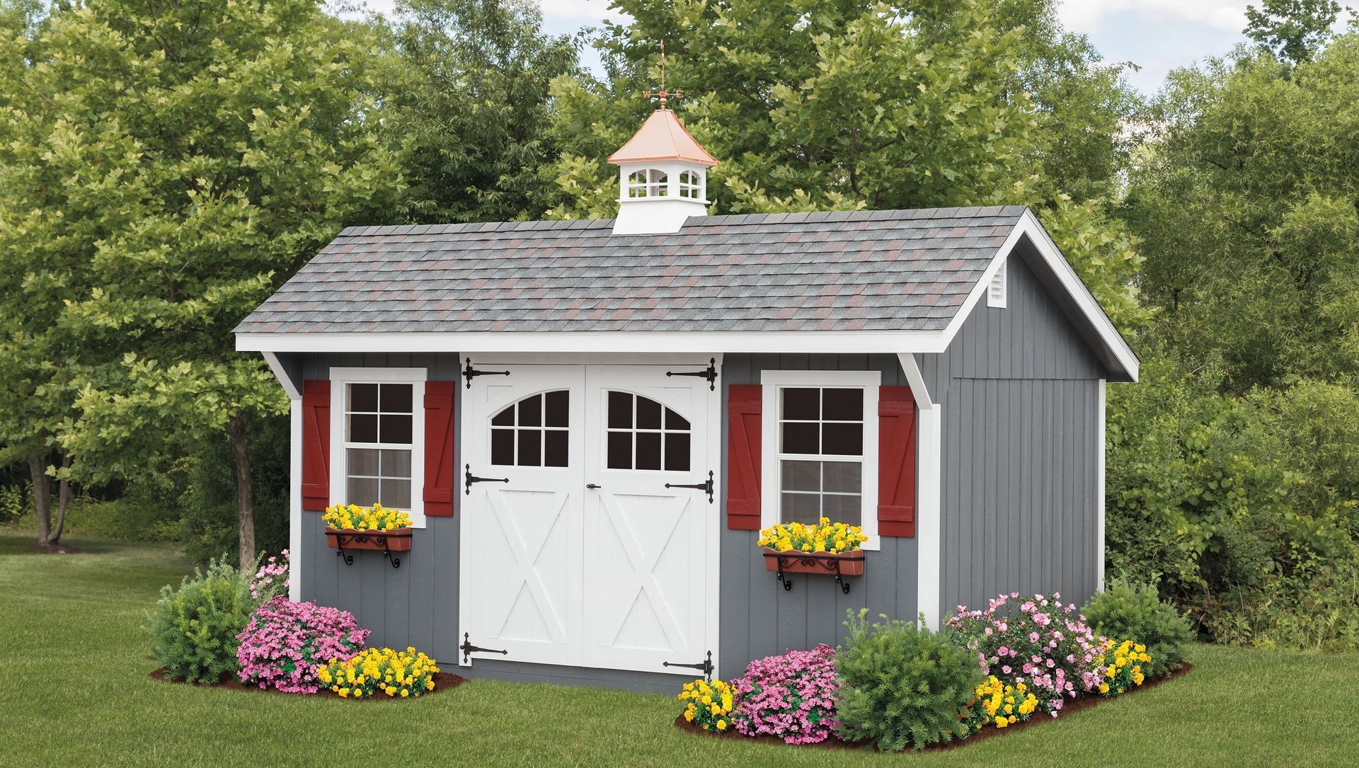 Small gray garden shed with white trim, red shutters, flower boxes, and blooming flowers in front.