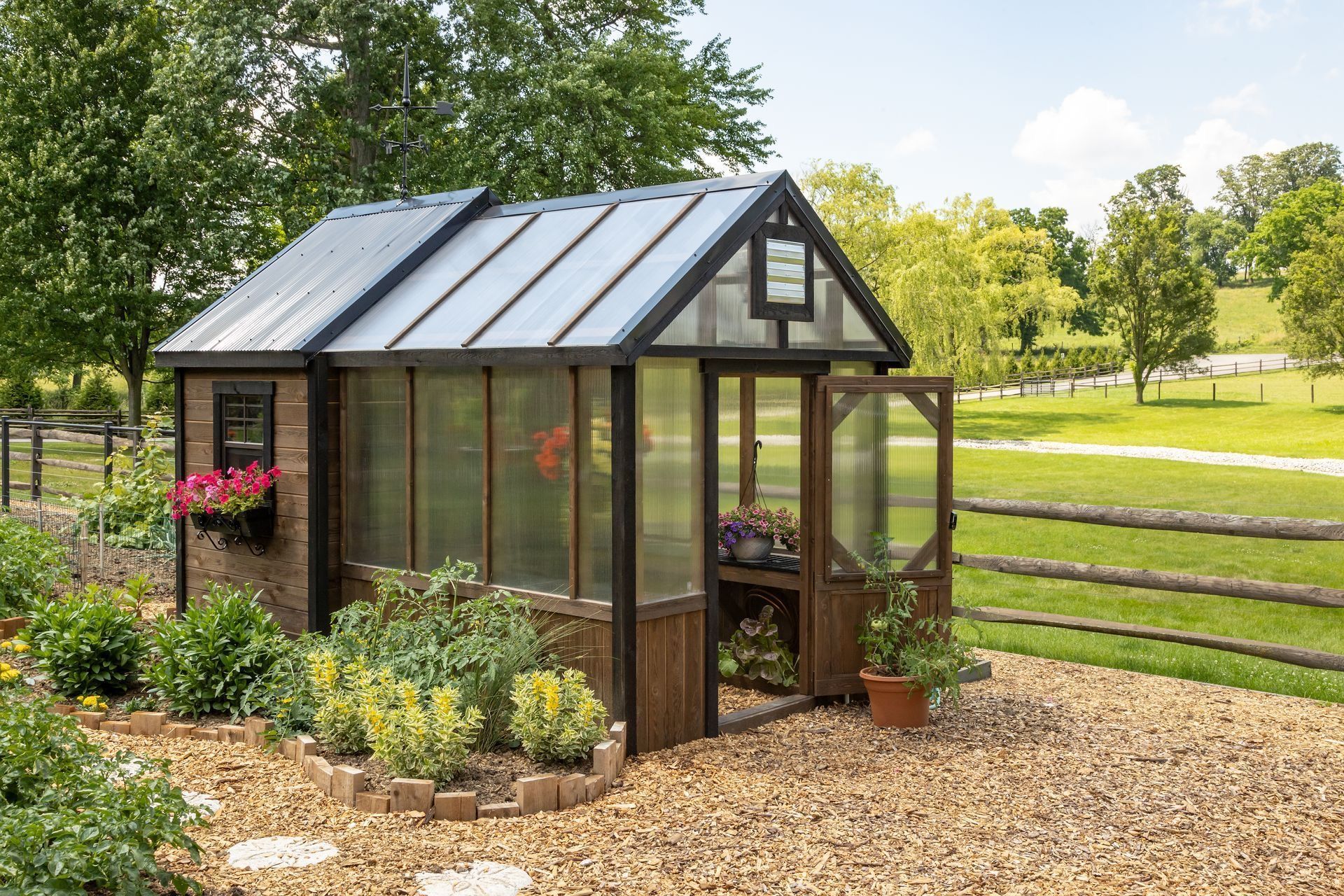 Small glass greenhouse in a garden, with raised beds, a gravel path, and a wooden fence.