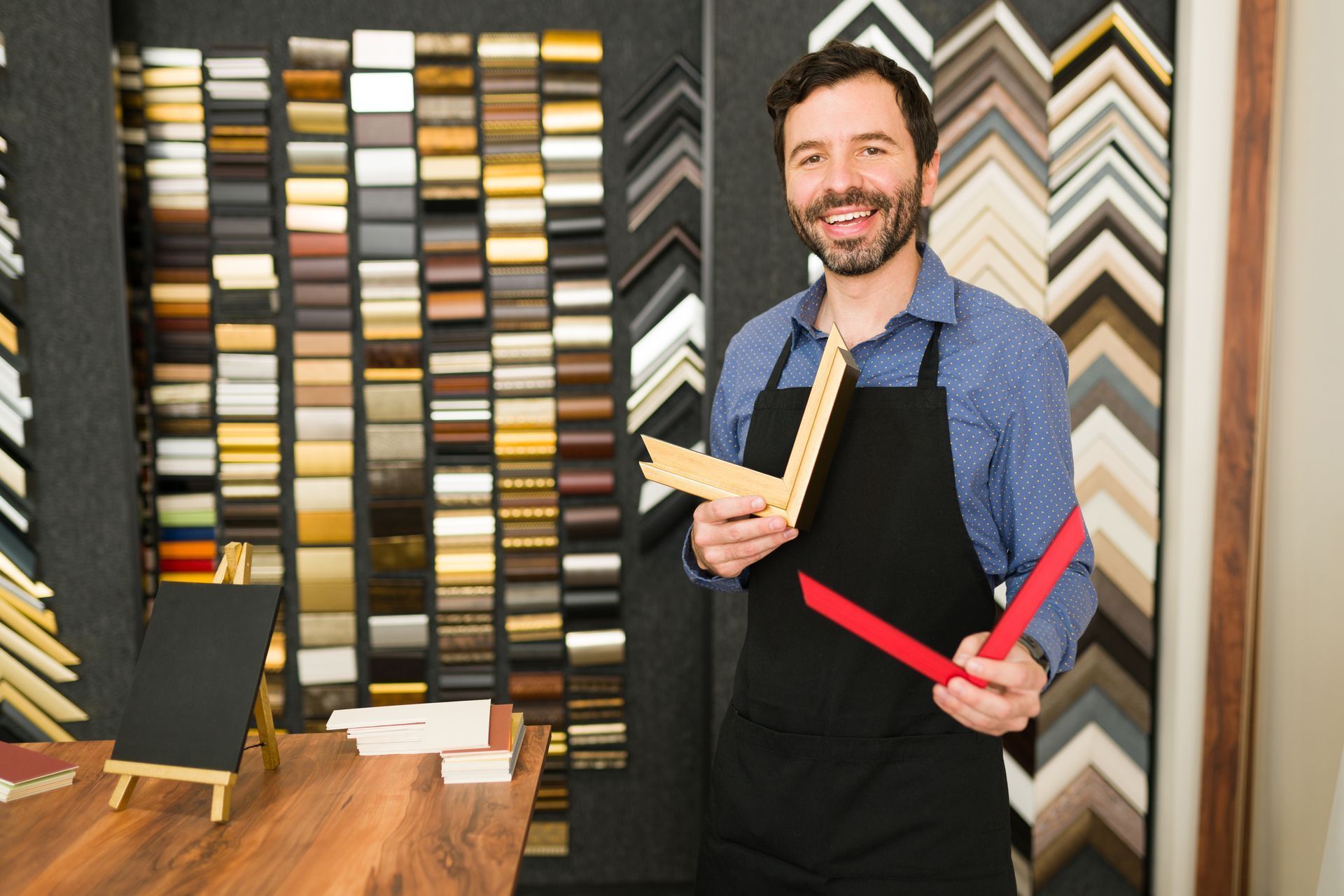 A smiling framer in a black apron holds sample picture frame corners against a wall filled with various molding options.
