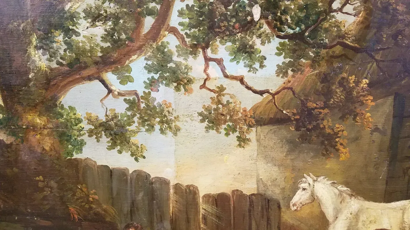 A white horse stands by a wooden fence and a stone building, sheltered beneath the branches of large, leafy trees.