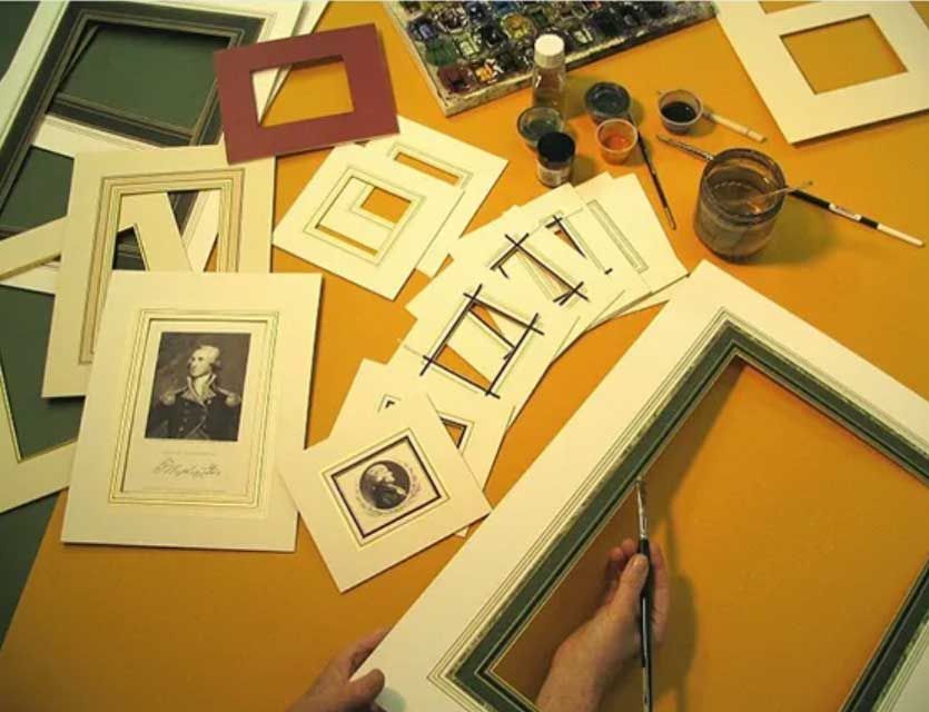 Hands paint decorative lines on a mat board, surrounded by framed prints, matting samples, and art supplies on a desk.