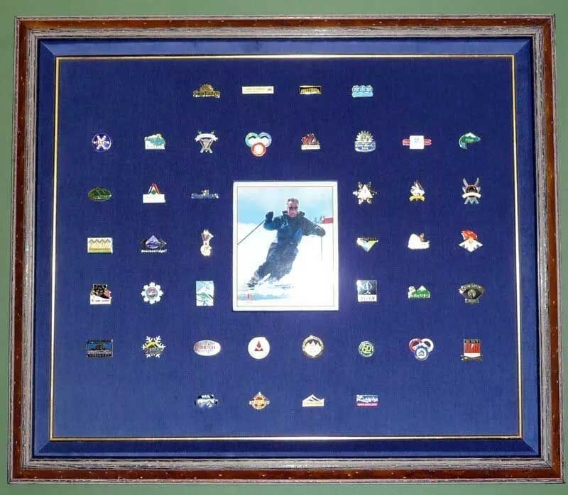 A framed blue display board featuring a central photo of a skier surrounded by many souvenir pins in a grid.